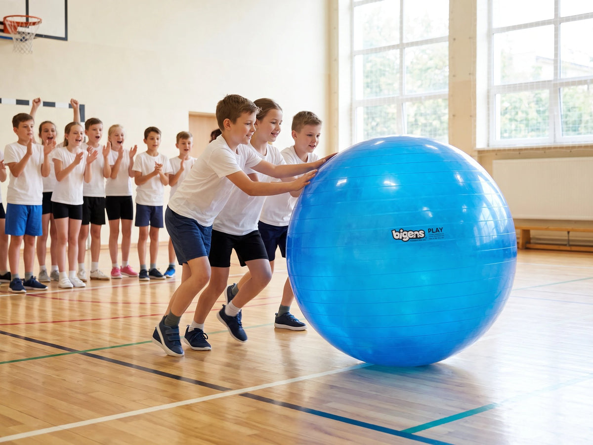 Three pupils pushing a giant blue Bigens Play Ball across a school gym while their class cheers