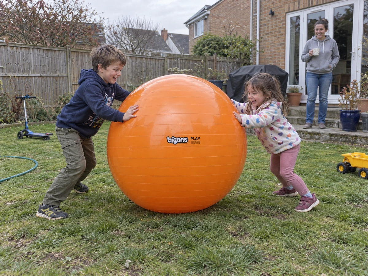 Two children pushing an orange Bigens Play Ball across a back garden lawn while a parent watches